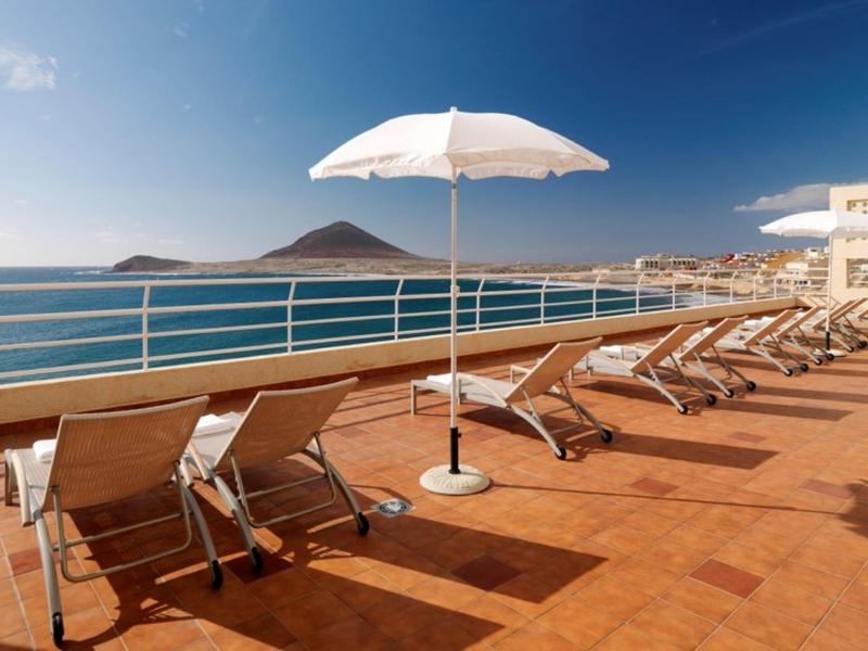 Terrazza soleggiata con lettini e ombrellone bianco con vista sul mare.