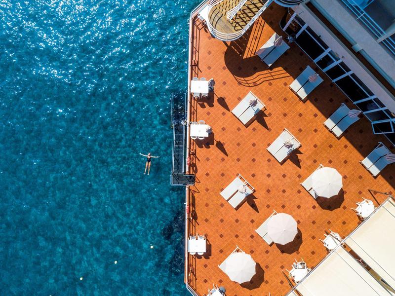 Vista dall'alto di una terrazza con tavoli, sedie e ombrelloni accanto all'acqua blu.