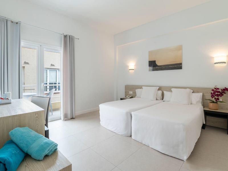 Camera d'hotel luminosa con due letti singoli, scrivania e balcone con luce naturale.