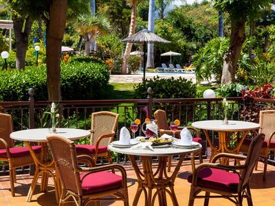 Area terrazza di un hotel con tavoli e sedie, circondata da piante tropicali e piscina sullo sfondo.