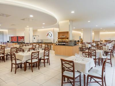 Salle à manger spacieuse d'hôtel avec tables blanches et chaises en bois sous un éclairage de plafond lumineux.