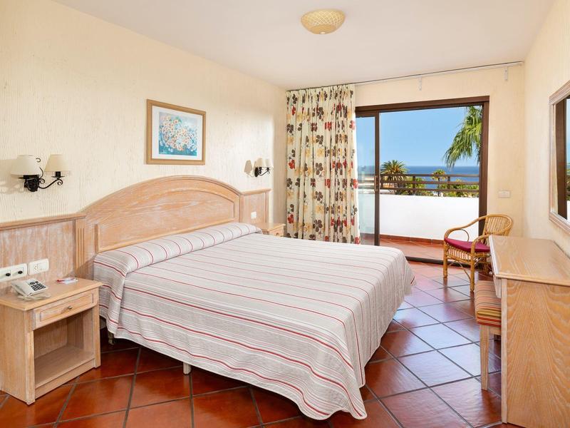 Jasný hotelový pokoj s velkou postelí, dřevěným nábytkem a výhledem na terasu a moře.