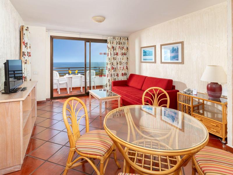 Interiér prázdninového pokoje s červenooranžovou pohovkou, malým jídelním stolem a výhledem na moře.