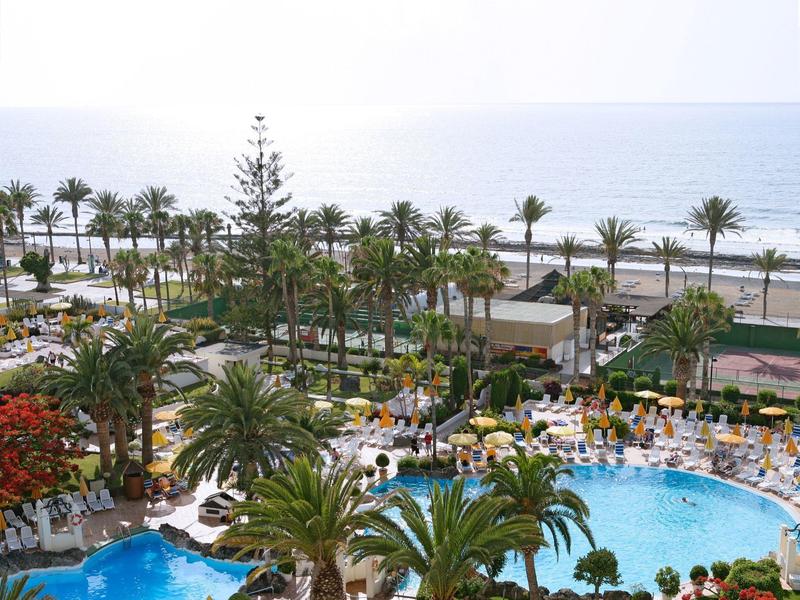 Ośrodek z dużym zakrzywionym basenem, palmami i morzem w tle pod jasnym niebem.