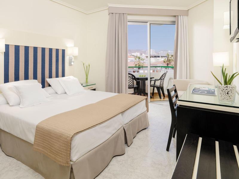 Jasny pokój hotelowy z dwoma łóżkami, beżowym wystrojem, biurkiem i balkonem z widokiem na miasto.