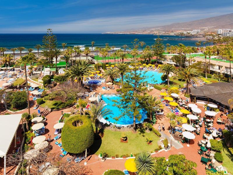 Ośrodek z basenami, parasolami, palmami i widokiem na ocean pod niebieskim niebem.