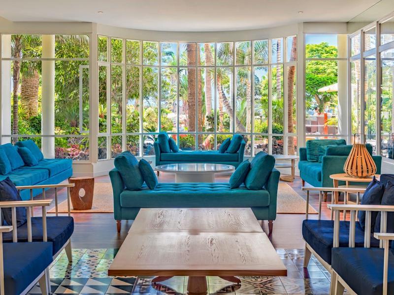 Jasny salon hotelowy z niebieskimi sofami, drewnianymi stołami i dużymi oknami z widokiem na ogród.