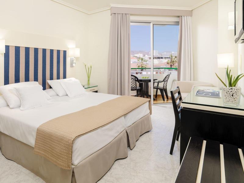 Jasny pokój hotelowy z podwójnym łóżkiem, biurkiem i balkonem z widokiem na miasto.