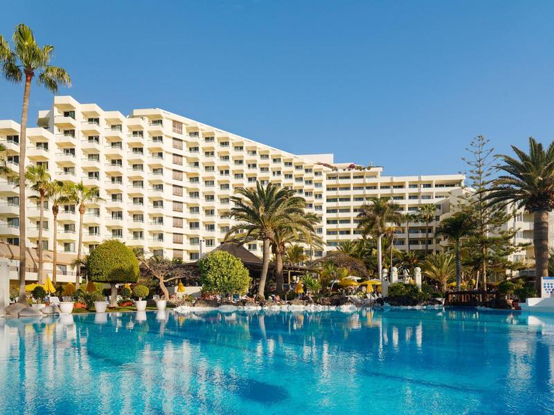 Duży hotel z zakrzywioną fasadą i palmami wokół jasnoniebieskiego basenu pod słonecznym niebem.