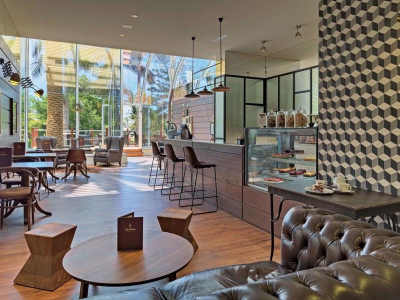 Nowoczesny salon hotelowy z skórzaną sofą, drewnianymi stolikami, barem i dużymi oknami wpuszczającymi światło.