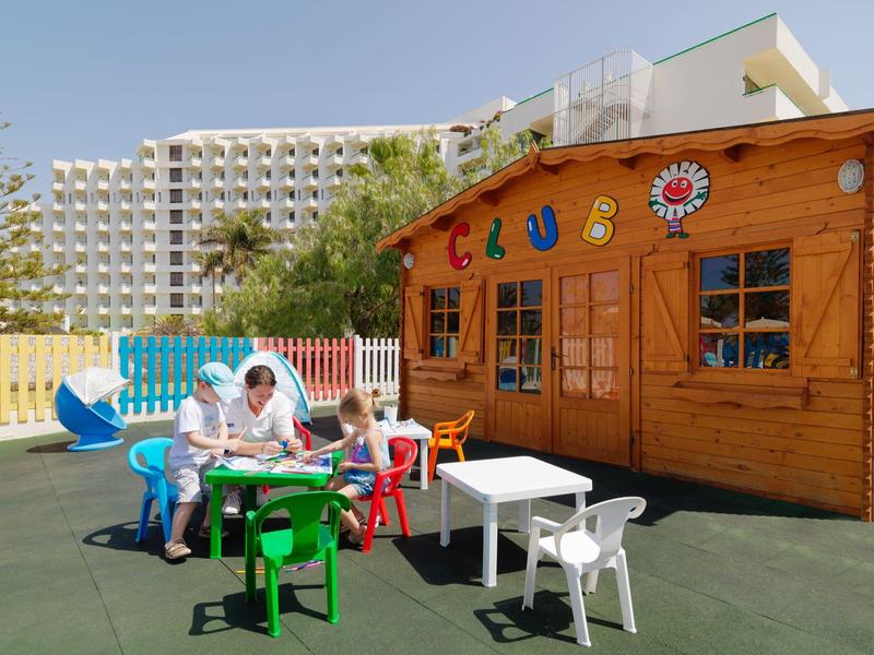 Dzieci bawiące się przed kolorowym drewnianym domkiem z zjeżdżalnią obok dużego hotelu.