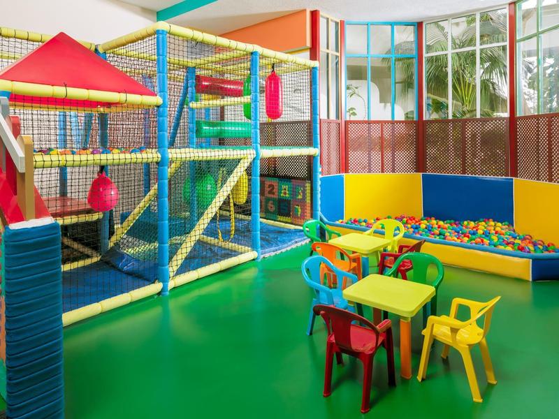 Wewnętrzny plac zabaw z kolorową konstrukcją do wspinaczki, basenem z piłeczkami i małymi stolikami z krzesłami.