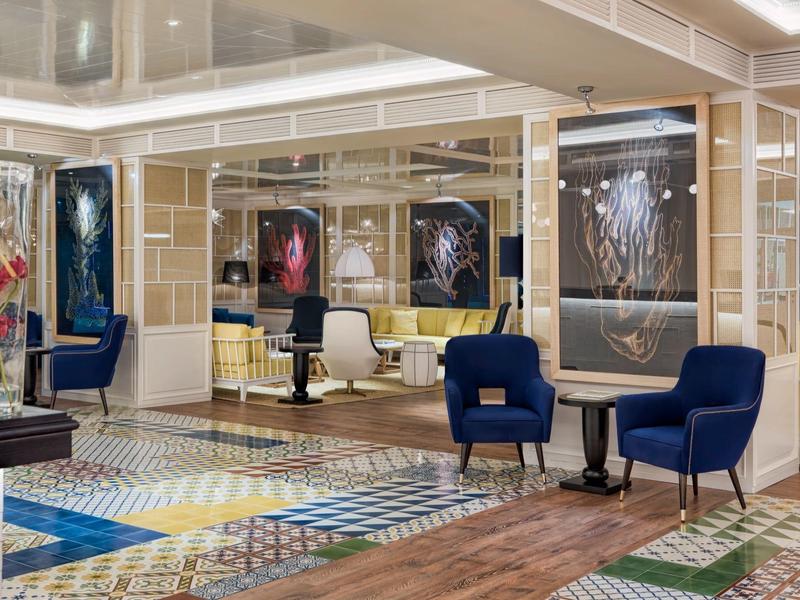 Nowoczesny hotelowy salon z kolorową podłogą, niebieskimi fotelami, szklanymi wystawami i dziełami sztuki na ścianach.