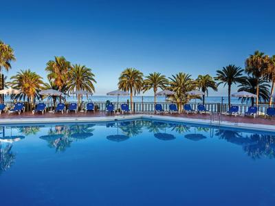 Piscina blu con sedie a sdraio e palme sotto un cielo sereno.