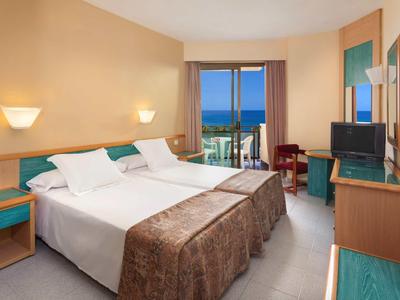 Helles Hotelzimmer mit Doppelbett, Balkon, Schreibtisch, Fernseher und Nachtlampen an beigefarbenen Wänden.