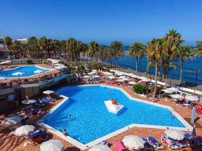 Hotel pool with sun umbrellas and palm trees by the sea under a clear blue sky.