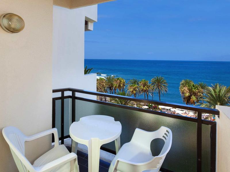Balcone con tavolo rotondo e sedie, vista sul mare e palme.