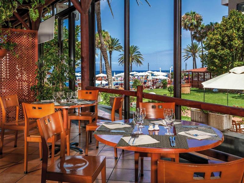 Terrazza di ristorante con vista su giardino e palme sotto un cielo azzurro.