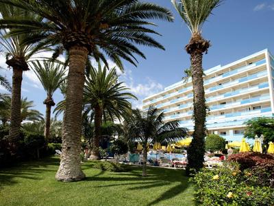 Modern hotel building with balconies surrounded by palm trees and a lush garden under a blue sky.