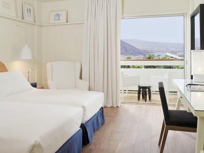 Bright hotel room with two beds, desk, chair, and balcony with mountain view.