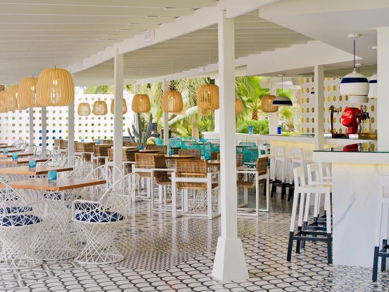 Bright open restaurant area with white columns, wicker chairs, and hanging lanterns by the beach.
