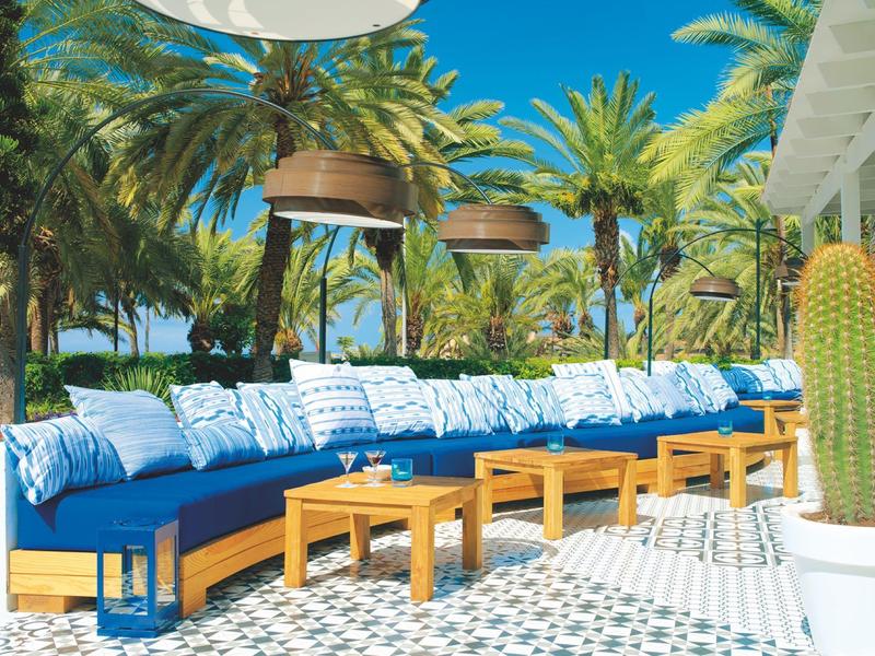 Outdoor seating area with blue cushions and wooden tables surrounded by palm trees.