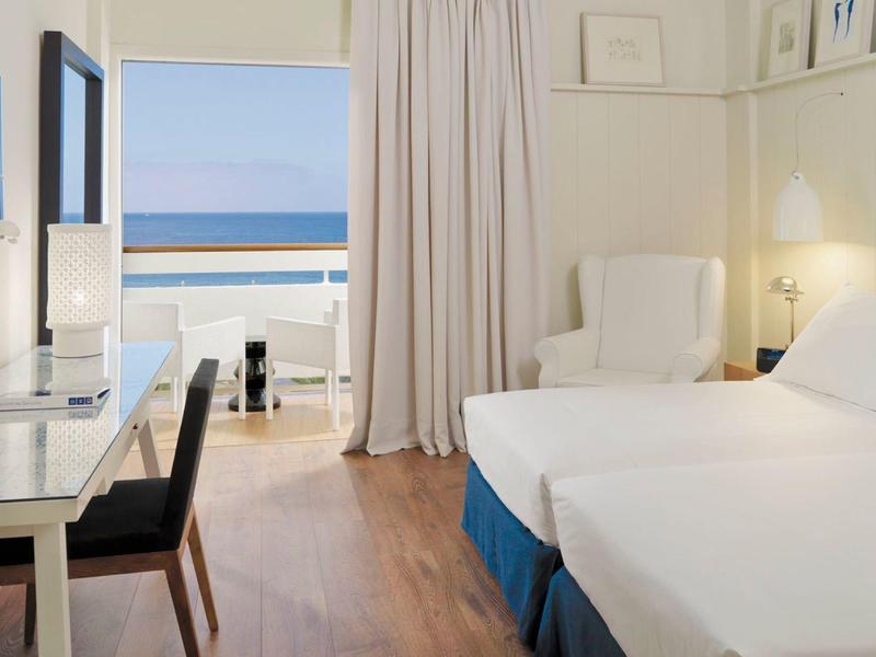 Cozy hotel room with twin beds, desk, and ocean view through balcony door.