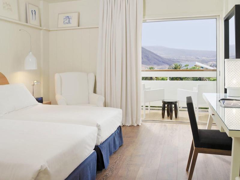 Bright hotel room with two beds, desk, chair, and balcony with mountain view.