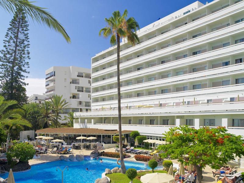 Modern hotel with outdoor pool, reclining chairs, palm trees, and clear blue sky.