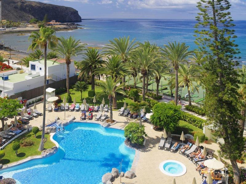 Resort pool area with palm trees, sun loungers, and ocean view under a partly cloudy sky.