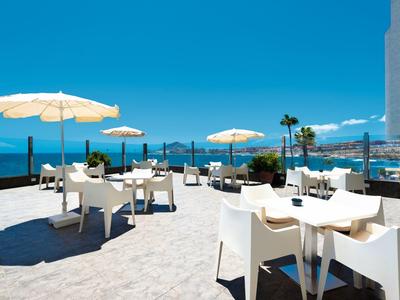 Taras z białymi stołami i krzesłami, parasolami i widokiem na niebieskie morze.