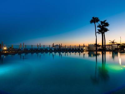 Vue du soir d'une piscine éclairée avec des palmiers et un port en arrière-plan.