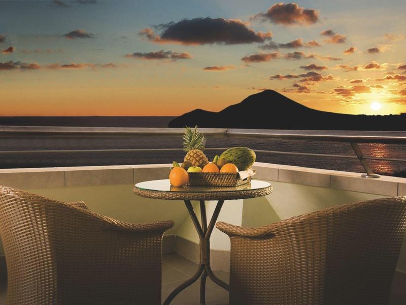 Zachód słońca nad morzem z tarasem, stołem i miską owoców na pierwszym planie.