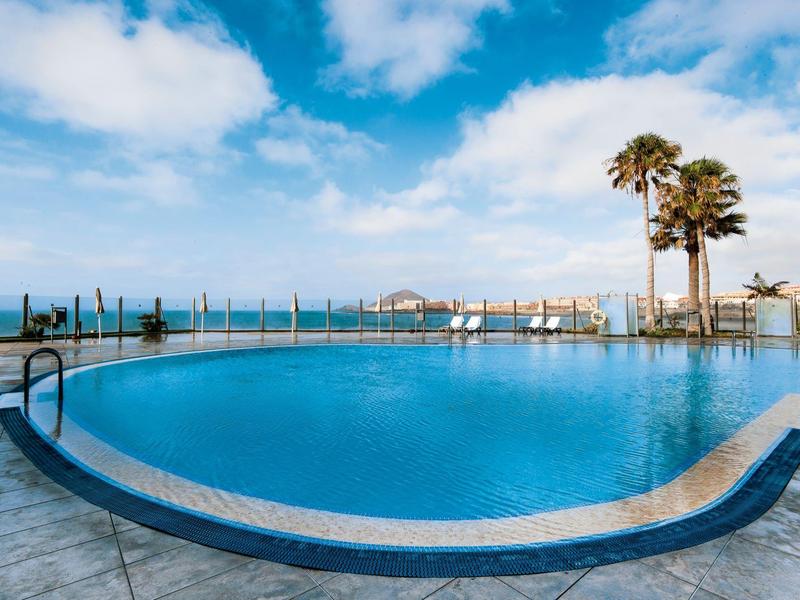 Okrągły basen hotelowy z widokiem na morze pod niebieskim niebem i palmami.