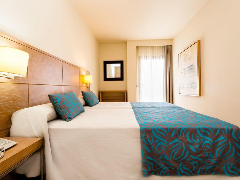 Jasny pokój hotelowy z dużym łóżkiem, drewnianym zagłówkiem i niebieskimi poduszkami w wzory.