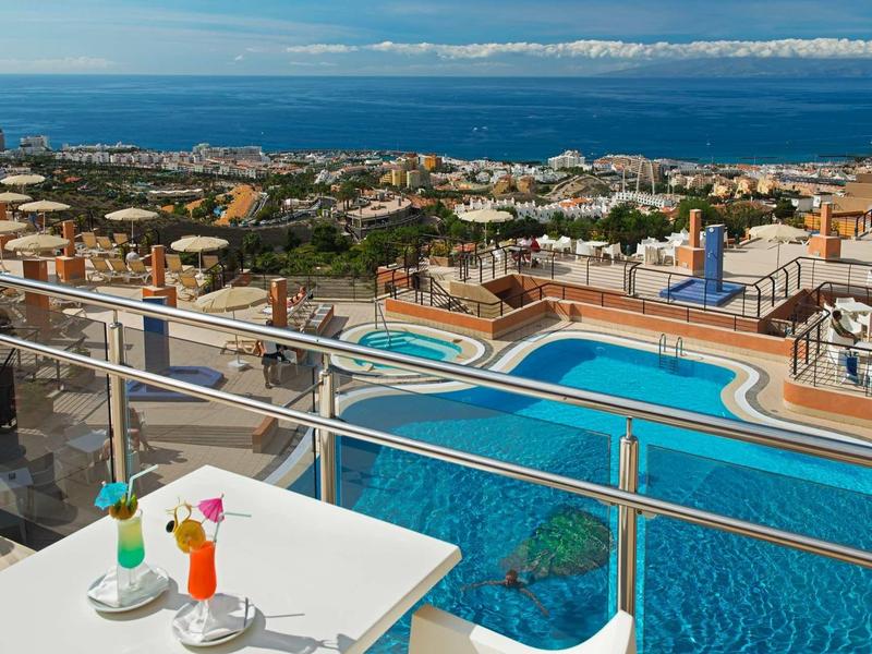 Blick auf Pool, Stadt und Meer mit blauem Himmel, Tisch mit bunten Cocktails auf Balkon im Vordergrund.