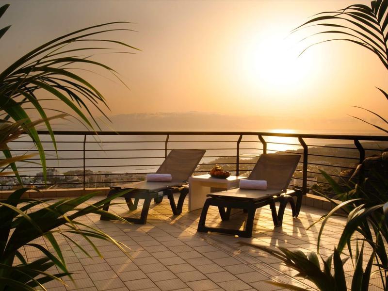 Zwei Liegestühle auf einer Terrasse mit Pflanzen, Blick auf Sonnenuntergang über dem Wasser.