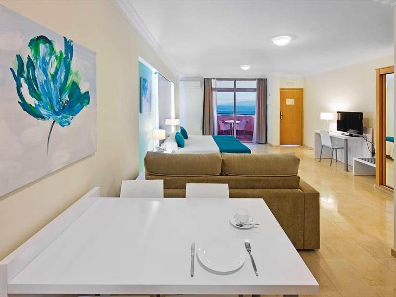 Moderner Wohnraum mit Essbereich, Sofa, Bett, Schreibtisch und großer Pflanzenmalerei an der Wand.