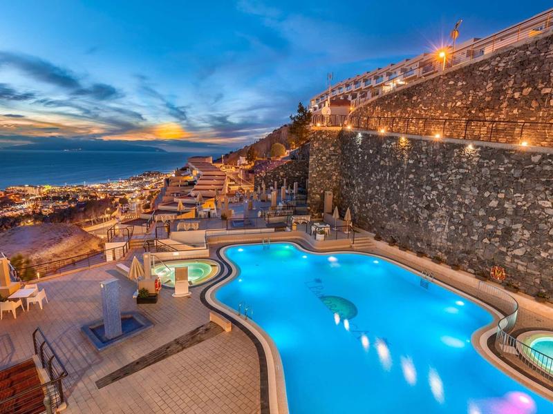 Beleuchteter Pool mit Liegestühlen neben einer hohen Steinmauer bei Sonnenuntergang.