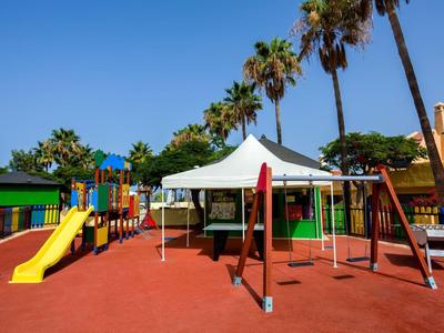 Area giochi colorata per bambini con scivoli, altalene e un tendone bianco sotto un cielo azzurro.