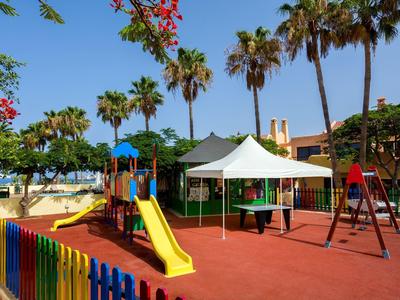 Parco giochi colorato con scivolo, altalene e tenda bianca sotto palme.