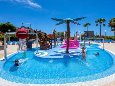 Piscina per bambini con scivolo e fontane d'acqua in un'area piscina soleggiata con palme.