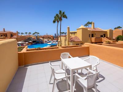 Terrazza con mobili da giardino bianchi e vista su piscina e palme in un villaggio turistico.