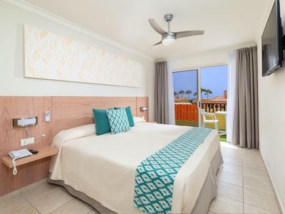 Camera d'hotel luminosa con letto matrimoniale, ventilatore a soffitto e balcone con vista sul mare.