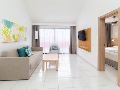 Soggiorno moderno con piastrelle chiare, divano, TV e accesso al balcone in una camera d'hotel.