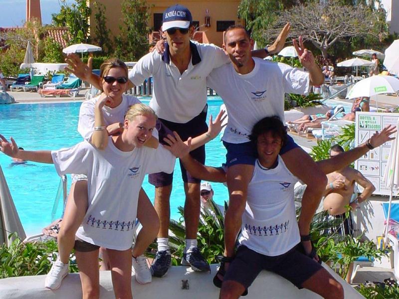 Cinque persone sorridenti in costume da bagno posano davanti a una piscina in una giornata di sole.