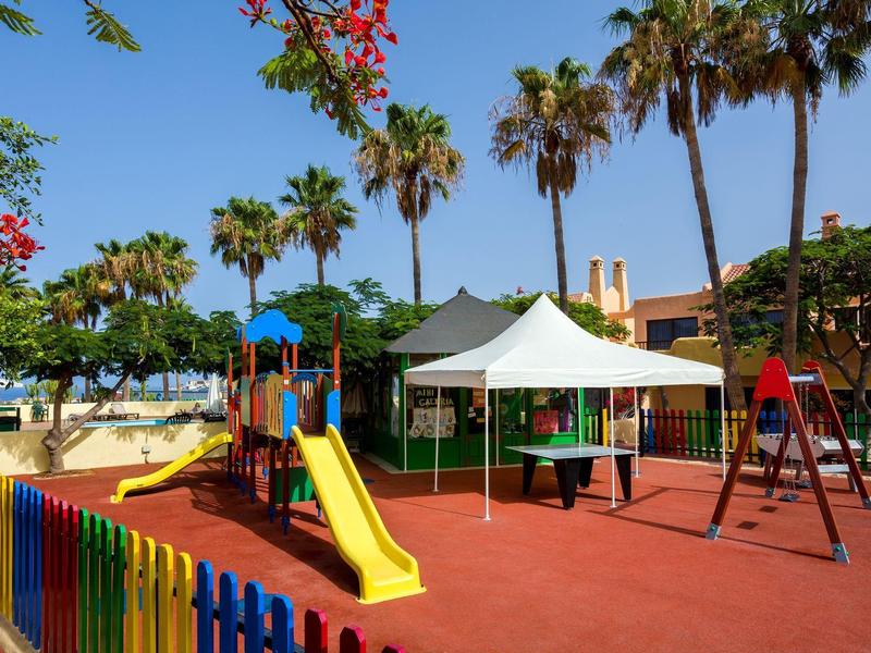 Parco giochi colorato con scivolo, altalene e tenda bianca sotto palme.