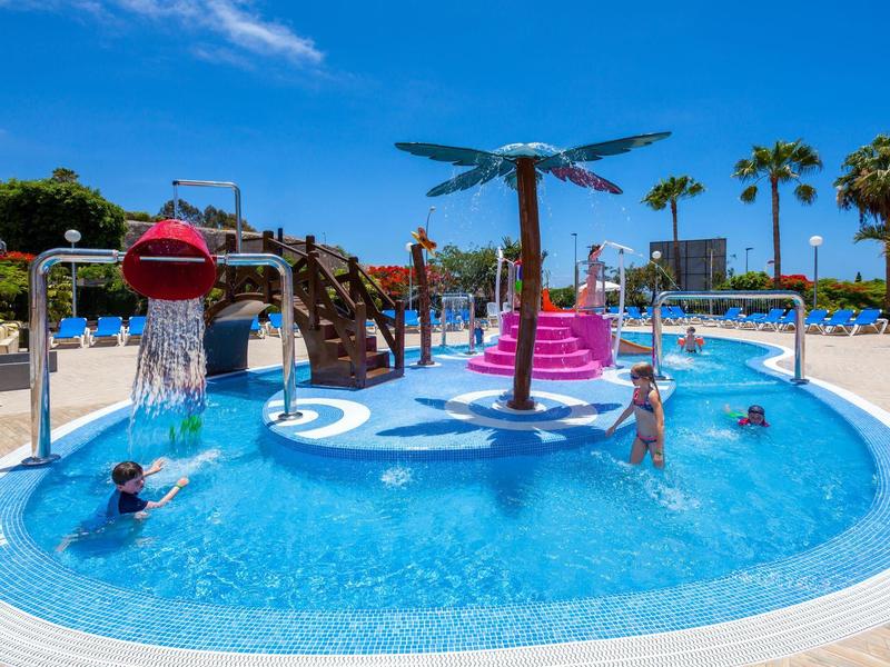 Piscina per bambini con scivolo e fontane d'acqua in un'area piscina soleggiata con palme.