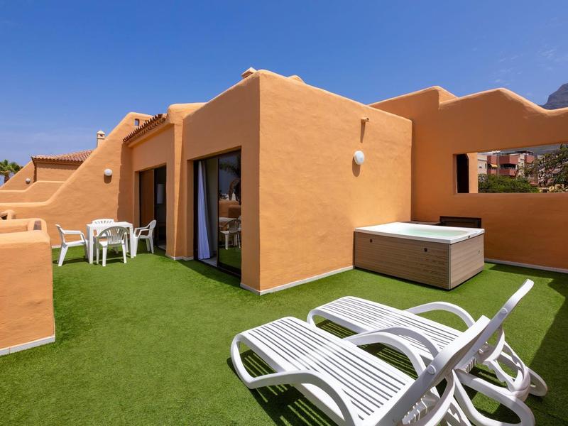 Terrazza con prato sintetico verde, lettini bianchi e vasca idromassaggio in un edificio alberghiero.