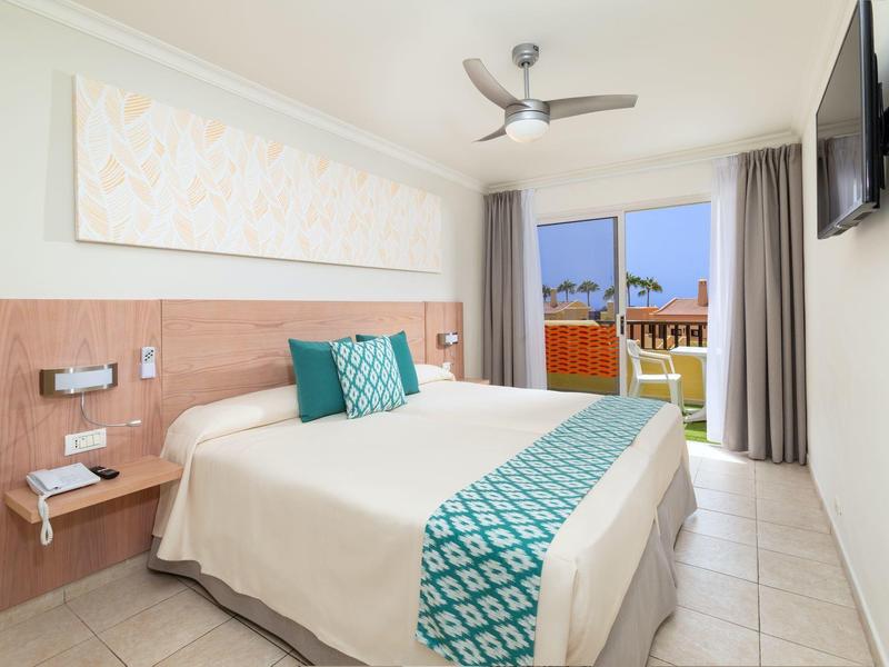 Camera d'hotel luminosa con letto matrimoniale, ventilatore a soffitto e balcone con vista sul mare.